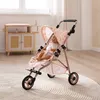 A doll is sitting in a pink doll stroller in a room with wooden furniture and a rug. Náhľad obrázku produktu