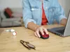 A person's hands are using a red and black wireless mouse on a wooden desk next to a laptop. There are also two tubes of paint and a pair of glasses on the desk. Anteprima dell’immagine prodotto