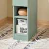 A close-up view of a narrow green nightstand with two shelves, a basket on the top shelf, and two boxes on the bottom shelf. The nightstand is placed on a patterned rug next to a wall. Náhľad obrázku produktu