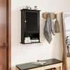 A black wall-mounted cabinet with a louvered door, placed on a wall above a bench. There are decorative items on top of the cabinet and items on the shelf below. A towel and a bag are hanging on a wooden coat rack to the right. Podgląd zdjęcia produktu