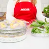 A person wearing a red apron with a logo is standing behind a food dehydrator with multiple trays. Some trays contain yellow slices of food, while another tray holds fresh green basil leaves. Náhľad obrázku produktu