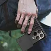 A person holding a phone case with a ring on the back, with a backpack in the background. Produktbild-Vorschau