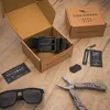 A black leather belt is displayed in its open packaging, alongside sunglasses, a multi-tool, and product tags, all arranged on a wooden surface. Produktbild-Vorschau