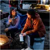 Two women sitting by a campfire, holding mugs, with a cooler and a vehicle in the background. Voir les images du produit