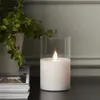 A white LED candle flickers inside a glass cylinder, placed on a dark surface next to a stack of magazines and a potted plant. Podgląd zdjęcia produktu