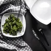 A square white plate of kale salad is placed on a black surface, next to a stack of white plates, a checkered cloth, and black cutlery. Náhľad obrázku produktu