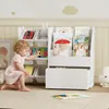 A young child is playing with a white bookshelf filled with books and toys in a nursery or playroom. Produktbild-Vorschau