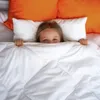 A young girl peeking out from under a white duvet in a bed with orange pillows. Náhľad obrázku produktu