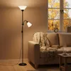 A floor lamp with a reading light is positioned next to a sofa in a living room with a window showing autumn trees. Produktbild-Vorschau