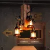 A rustic chandelier with four lantern-style lights hangs above a wooden table in a dimly lit room with brick and concrete walls. Náhled obrázku produktu