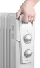 A hand is shown gripping the handle of a white oil-filled radiator heater. Náhled obrázku produktu