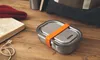 A stainless steel lunch box with an orange strap sits on a wooden table next to an open book and a red knitted hat. Podgląd zdjęcia produktu