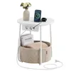 A white round side table with integrated power outlets and USB ports, featuring a phone and smartwatch charging, a vase with flowers, and storage for books and a blanket. Produktbild-Vorschau