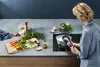A woman is cooking in a modern kitchen, using a pressure cooker on an induction stovetop. Fresh vegetables are prepared on a cutting board. Náhled obrázku produktu