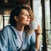 A woman holding a coffee cup indoors, looking out a window. Produktbild-Vorschau