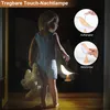 A young child holding a teddy bear and a bird-shaped night light in a dimly lit room. Produktbild-Vorschau