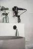 A black hairdryer is mounted on a wall rack above a white sink, with a green plant in a vase and a green towel hanging from a hook. Produktbild-Vorschau
