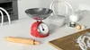 A kitchen counter with baking ingredients and tools, including a red analog kitchen scale, a rolling pin, flour, eggs, and milk. Náhľad obrázku produktu