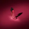 A high-heeled shoe-shaped perfume bottle with a glittery red finish and a black cap, casting a shadow on a deep pink background. Anteprima dell’immagine prodotto