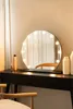 A round vanity mirror with lights is placed on a black dresser, with a chair in the foreground. Produktbild-Vorschau