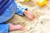 A child's bare feet and legs in blue jeans are in the sand, with a yellow toy bucket in the background. Podgląd zdjęcia produktu