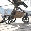 A close-up shot of a stroller's wheels and storage basket, with a person's leg visible on the left, on a wooden boardwalk. Náhled obrázku produktu