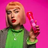 A woman with pink hair and bold makeup holds a pink bottle of Tigi Bed Head After Party Super Smoothing Cream against a bright pink background. Podgląd zdjęcia produktu