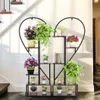 A heart-shaped plant stand with multiple shelves filled with potted plants, placed near a staircase. Produktbild-Vorschau