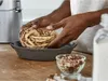 Hands are shaping a braided dough with chocolate filling into a pan, with a mixer and a bowl of nuts in the background. Voir les images du produit
