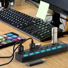A USB hub with multiple ports and switches is connected to a smartphone and a USB drive, with a keyboard and a computer monitor in the background. Produktbild-Vorschau