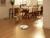 A white robot vacuum cleaner is cleaning a wooden floor in a dining room, with a dog nearby. Produktbild-Vorschau