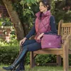 A woman sitting on a wooden bench in a garden setting, wearing equestrian-style clothing and holding a matching bag. Podgląd zdjęcia produktu