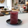 A lit burgundy pillar candle sits on a black plate on a white table, with a blurred living room scene in the background. Anteprima dell’immagine prodotto