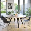 A round wooden dining table with black metal legs, surrounded by black chairs in a brightly lit room with large windows overlooking a garden. Anteprima dell’immagine prodotto