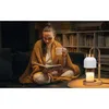 A woman is sitting on a couch, wrapped in a blanket, reading a book and holding a mug. A candle warmer lamp is on a side table next to her. Náhled obrázku produktu