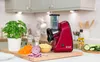 A red Russell Hobbs food processor is displayed on a wooden cutting board with various ingredients, including shredded carrots, red onion, grated cheese, and cucumber slices, in a kitchen setting. Produktbild-Vorschau
