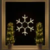 A snowflake-shaped LED light decoration hangs in a window at night, flanked by two potted evergreen trees decorated with string lights. Produktbild-Vorschau
