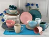 A collection of ceramic tableware, including plates, bowls, and mugs, arranged on a blue placemat with blue flowers in the background. Náhľad obrázku produktu