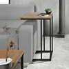 A narrow side table with a wooden top and black metal legs is placed next to a gray sofa. A small plant in a black pot and a book are on the table. A vase with dried flowers is on a lower table in the foreground. Produktbild-Vorschau