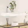 A white floating shelf with a drawer is mounted on a white wall. A framed abstract artwork and a glass vase with green leaves are placed on the shelf. Below the shelf, on a wooden surface, is a glass bottle with reeds and a stack of books. Produktbild-Vorschau