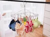 A metal sock dryer with multiple clips is hanging from a rod, with various items of underwear and a bra attached to it. The background shows a window with sheer curtains and a white dresser. Produktbild-Vorschau