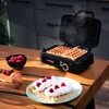 A waffle maker with two waffles cooking inside is shown on a wooden table. Nearby, there is a bowl of raspberries, two eggs, a honey dipper, and a plate with two finished waffles topped with cream and raspberries. Produktbild-Vorschau