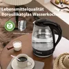 A glass electric kettle is shown on a round wooden table, next to a cup of coffee and a small pitcher of milk. The background suggests a cozy indoor setting with a lamp and textured rug. Produktbild-Vorschau