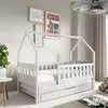A white wooden children's house bed with a pull-out drawer, shown in a bedroom setting with artwork on the wall and a plant. Produktbild-Vorschau