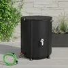 A black foldable water barrel with a green hose and red valve attachment sits on a light gray patio next to a stone wall and green foliage. Náhled obrázku produktu