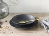 A dark gray stoneware bowl and plate set with a gold spoon resting on top, arranged on a wooden surface with a glass vase and dried branches in the background. Produktbild-Vorschau