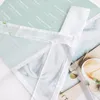 A close-up view of a light green fabric with white text and a white ribbon tied in a cross shape. Produktbild-Vorschau
