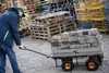 A person is pulling a cart loaded with paving stones in an outdoor storage area with stacks of pallets and metal cages in the background. Produktbild-Vorschau