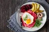 A plate of food with a fried egg, mashed beets, sliced beets, pickles, and herring, served on a wooden table. Produktbild-Vorschau