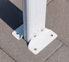 A close-up view of the base of a white metal post, secured to a paved surface with metal brackets and screws. Podgląd zdjęcia produktu